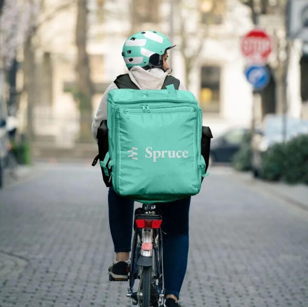 A bicycle delivery rider with a teal backpack and helmet branded with Spruce