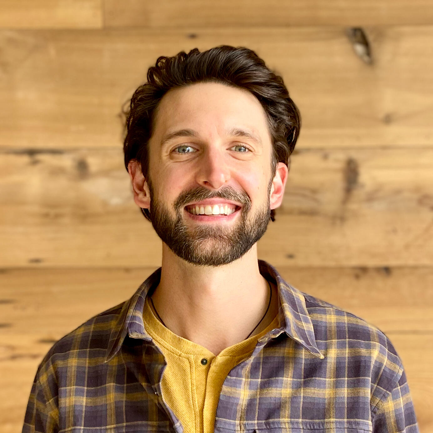 Devon de Balasi Brown, smiling man with plaid shirt in front of wooden background
